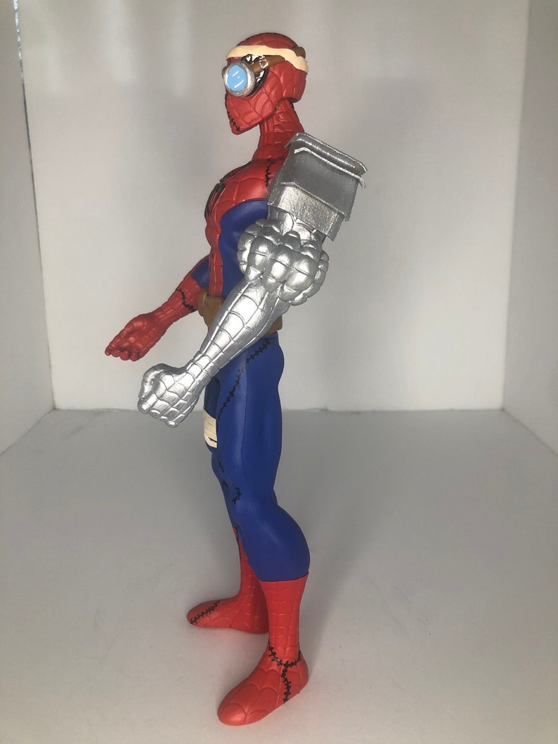 May include: A Spider-Man action figure with a steampunk-inspired design. The figure features a red and blue suit, a silver robotic arm, and goggles. The figure is approximately 12 inches tall.