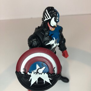 Puede incluir: Figura de acci&oacute;n negra y azul de un superh&eacute;roe sosteniendo un escudo. La figura tiene una cara blanca con una lengua roja y detalles blancos. El escudo es rojo, blanco y azul con una estrella blanca.