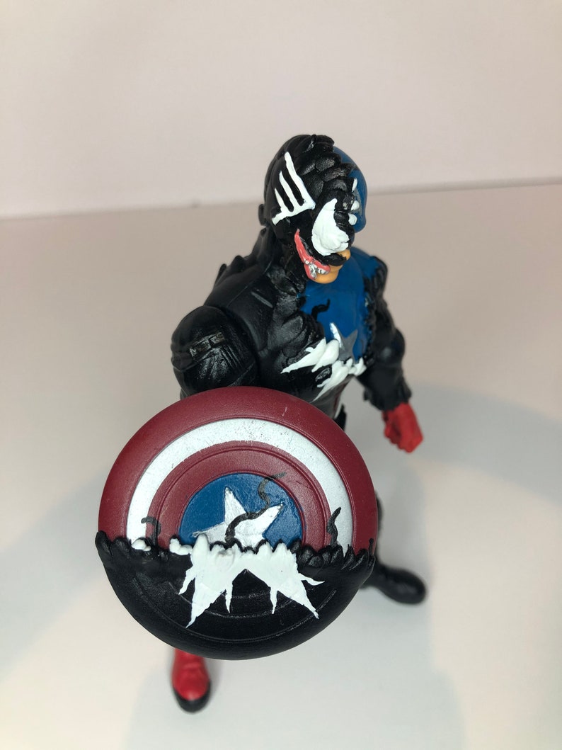 Puede incluir: Figura de acci&oacute;n negra y azul de un superh&eacute;roe sosteniendo un escudo. La figura tiene una cara blanca con una lengua roja y detalles blancos. El escudo es rojo, blanco y azul con una estrella blanca.