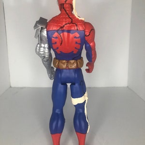 May include: A Spider-Man action figure, viewed from the back, featuring a red and blue costume with stitched details. The figure has a silver robotic arm and bandages around the head and leg. The figure has a brown utility belt.