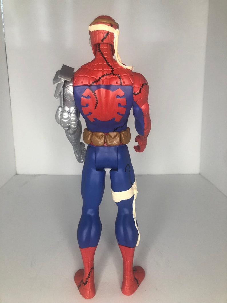May include: A Spider-Man action figure, viewed from the back, featuring a red and blue costume with stitched details. The figure has a silver robotic arm and bandages around the head and leg. The figure has a brown utility belt.