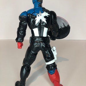 Puede incluir: Figura de acci&oacute;n del Capit&aacute;n Am&eacute;rica, vista desde atr&aacute;s, con la cabeza azul y un emblema de estrella en blanco y negro en la espalda. La figura lleva un traje negro con guantes rojos y una pierna roja y azul. Un escudo se sostiene en el brazo derecho.