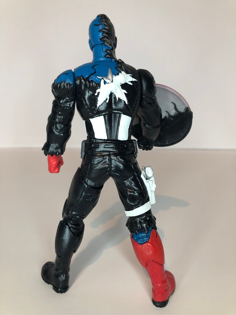 Puede incluir: Figura de acci&oacute;n del Capit&aacute;n Am&eacute;rica, vista desde atr&aacute;s, con la cabeza azul y un emblema de estrella en blanco y negro en la espalda. La figura lleva un traje negro con guantes rojos y una pierna roja y azul. Un escudo se sostiene en el brazo derecho.