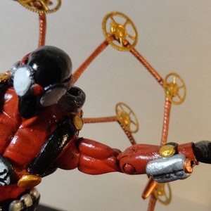 May include: A steampunk-inspired figurine with a red and black body, gold accents, and a black head. The figure has a spider emblem and is adorned with gear-like elements extending from its back.