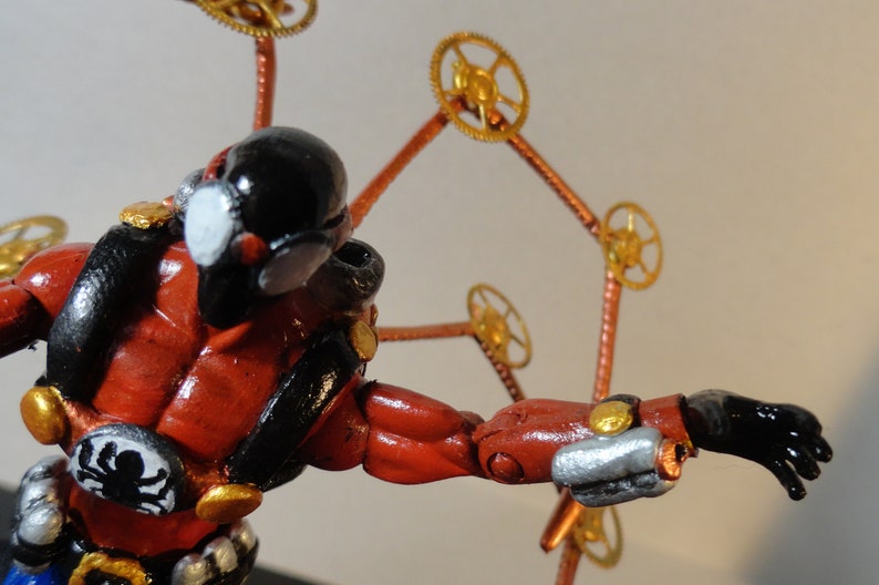 May include: A steampunk-inspired figurine with a red and black body, gold accents, and a black head. The figure has a spider emblem and is adorned with gear-like elements extending from its back.