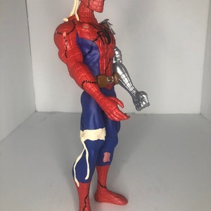 May include: A red, blue, and silver Spider-Man action figure with a unique design. The figure features a robotic arm, stitched seams, and bandages. The figure has a brown belt and a black spider emblem on the chest.