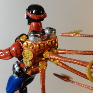 May include: A handcrafted figurine of a figure in a red and black suit with blue pants. The figure has metallic accents and copper-colored tubes and gears extending from its back, suggesting a steampunk aesthetic.