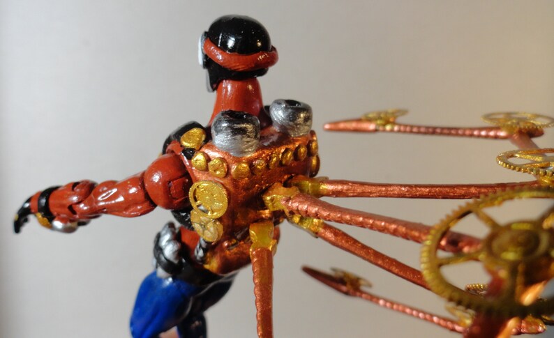 May include: A handcrafted figurine of a figure in a red and black suit with blue pants. The figure has metallic accents and copper-colored tubes and gears extending from its back, suggesting a steampunk aesthetic.