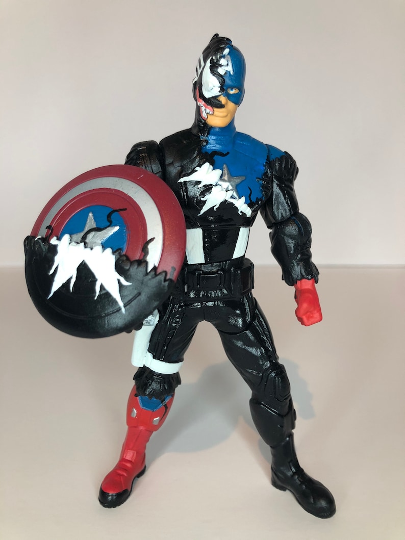 Puede incluir: Una figura de acci&oacute;n del Capit&aacute;n Am&eacute;rica con un dise&ntilde;o de simbionte Venom. La figura es predominantemente negra y azul, con un escudo rojo, blanco y azul. La cara de la figura est&aacute; dividida, un lado Capit&aacute;n Am&eacute;rica y el otro Venom.