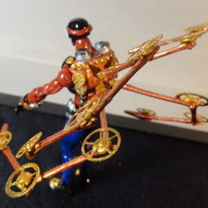 May include: A steampunk-inspired figurine with a red and copper-colored body, blue pants, and a black helmet. The figure has multiple copper arms with gold-colored gears attached, creating a mechanical wing-like structure.