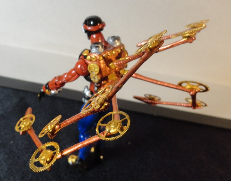 May include: A steampunk-inspired figurine with a red and copper-colored body, blue pants, and a black helmet. The figure has multiple copper arms with gold-colored gears attached, creating a mechanical wing-like structure.