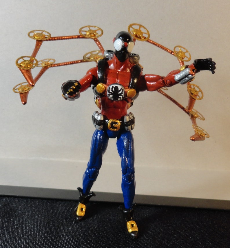 May include: A red, black, and blue action figure with gold clockwork-style appendages. The figure has a spider emblem on the chest and is posed with arms outstretched. The figure's legs are blue, and the boots have gold accents.