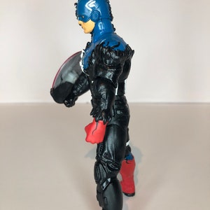 Puede incluir: Figura de acci&oacute;n de Capit&aacute;n Am&eacute;rica de perfil. La figura lleva un casco azul con una "A" blanca, un traje negro y azul, y sostiene un escudo. Las botas son negras y rojas. Art&iacute;culo de coleccionista.