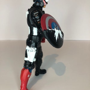 Puede incluir: Figura de acci&oacute;n en blanco y negro de un personaje con un escudo. La figura tiene un traje negro con detalles en blanco, una bota roja y blanca y un escudo con colores rojo, blanco y azul.