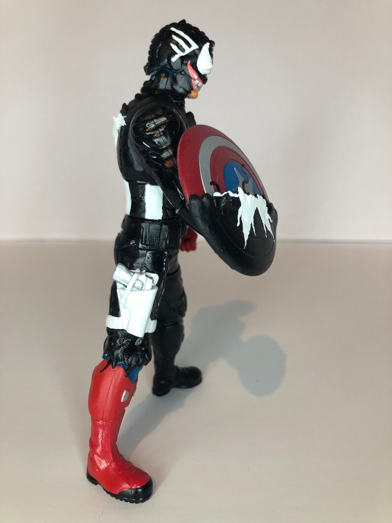 Puede incluir: Figura de acci&oacute;n en blanco y negro de un personaje con un escudo. La figura tiene un traje negro con detalles en blanco, una bota roja y blanca y un escudo con colores rojo, blanco y azul.