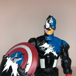 Puede incluir: Una figura de acci&oacute;n de Capit&aacute;n Am&eacute;rica con un dise&ntilde;o inspirado en Venom. La figura es principalmente azul y negra, sosteniendo un escudo rojo, blanco y azul. La cara y partes del cuerpo est&aacute;n divididas entre Capit&aacute;n Am&eacute;rica y Venom.