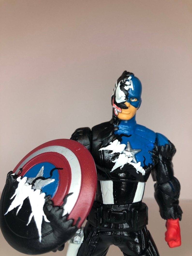 Puede incluir: Una figura de acci&oacute;n de Capit&aacute;n Am&eacute;rica con un dise&ntilde;o inspirado en Venom. La figura es principalmente azul y negra, sosteniendo un escudo rojo, blanco y azul. La cara y partes del cuerpo est&aacute;n divididas entre Capit&aacute;n Am&eacute;rica y Venom.