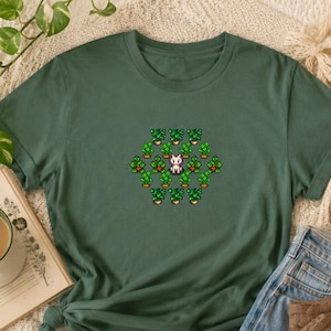 Könnte beinhalten: Ein dunkelolivgrünes T-Shirt mit einer zentralen Pixel-Art-Grafik. Das Design zeigt eine kleine weiße Katzenfigur, umgeben von einem sich wiederholenden Muster aus grünen Topfpflanzen mit orangefarbenen Akzenten, die in einer hexagonalen Gruppe angeordnet sind.