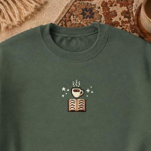 May include: A forest green crewneck sweatshirt featuring a small, centred pixel art graphic. The design depicts an open book with a steaming cup of coffee resting above it, surrounded by several small, white four-pointed star icons.