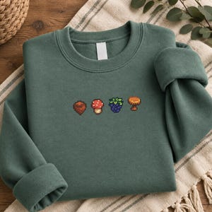 Könnte beinhalten: Ein waldgrünes Langarm-Sweatshirt mit einer kleinen, mittig platzierten Pixel-Art-Stickerei. Das Design besteht aus vier verschiedenen Symbolen: einer braunen Eichel, einem rot-weiß gepunkteten Pilz, einer dunkelvioletten Brombeere und einem beigen Pilz.