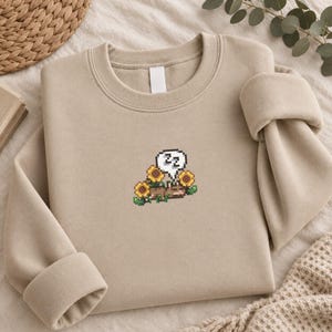Könnte beinhalten: Ein beigefarbenes Sweatshirt mit Rundhalsausschnitt und einer kleinen Pixel-Art-Stickerei auf der Brust. Das Motiv zeigt ein braunes Tier, das zwischen gelben Sonnenblumen mit grünen Blättern schläft, sowie eine weiße Sprechblase mit dem schwarzen Text ZZZ.