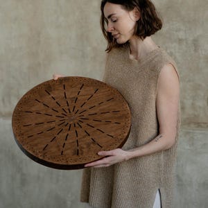 May include: A circular wooden board featuring a dark brown finish with a sunburst pattern of etched lines radiating from the center. The outer edge of the board is decorated with a repeating geometric triangle border pattern carved into the wood.