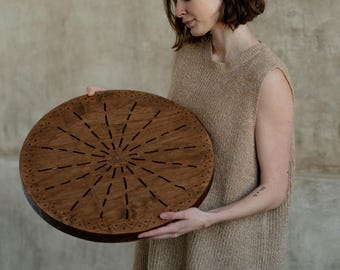May include: A circular wooden board featuring a dark brown finish with a sunburst pattern of etched lines radiating from the center. The outer edge of the board is decorated with a repeating geometric triangle border pattern carved into the wood.