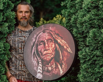 Puede incluir: Un hombre con barba sostiene un tambor redondo con una imagen pintada de un hombre nativo americano con un tocado de plumas. El tambor es marrón con acentos rojos y blancos.