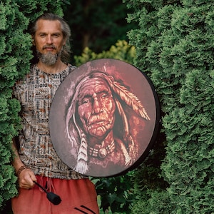 May include: A man with a beard holds a round drum with a painted image of a Native American man with a feather headdress. The drum is brown with red and white accents.