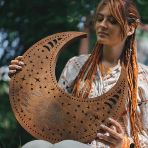 May include: A crescent moon-shaped wooden wall hanging with star cutouts and etched celestial designs. The brown-toned decor piece is held in front of a blurred green background.