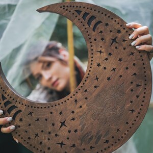 May include: A crescent moon-shaped wooden wall hanging with a dark brown finish. The moon features cut-out stars, a decorative owl design, and a series of small holes along the edge. The design is held by a person.