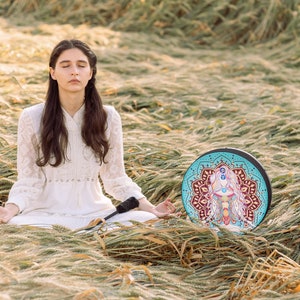 May include: A woman sits in a field of long grass, meditating with her eyes closed. She is wearing a white dress and has long dark hair. A round, colourful mandala drum sits in front of her.
