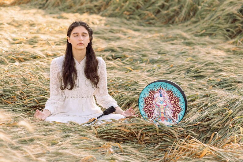 May include: A woman sits in a field of long grass, meditating with her eyes closed. She is wearing a white dress and has long dark hair. A round, colourful mandala drum sits in front of her.