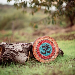 May include: A round wooden drum with a colourful mandala design and the symbol "Om" in the centre. The drum is resting on a tree log in a grassy area.