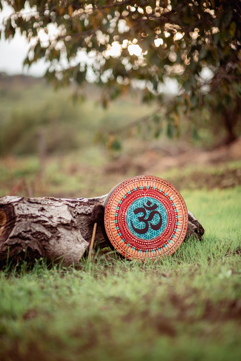 May include: A round wooden drum with a colourful mandala design and the symbol "Om" in the centre. The drum is resting on a tree log in a grassy area.
