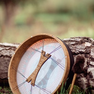 May include: A wooden frame drum with a white and blue patterned membrane and a wooden handle. The drum is resting on a tree trunk with a brown and black drum stick beside it.