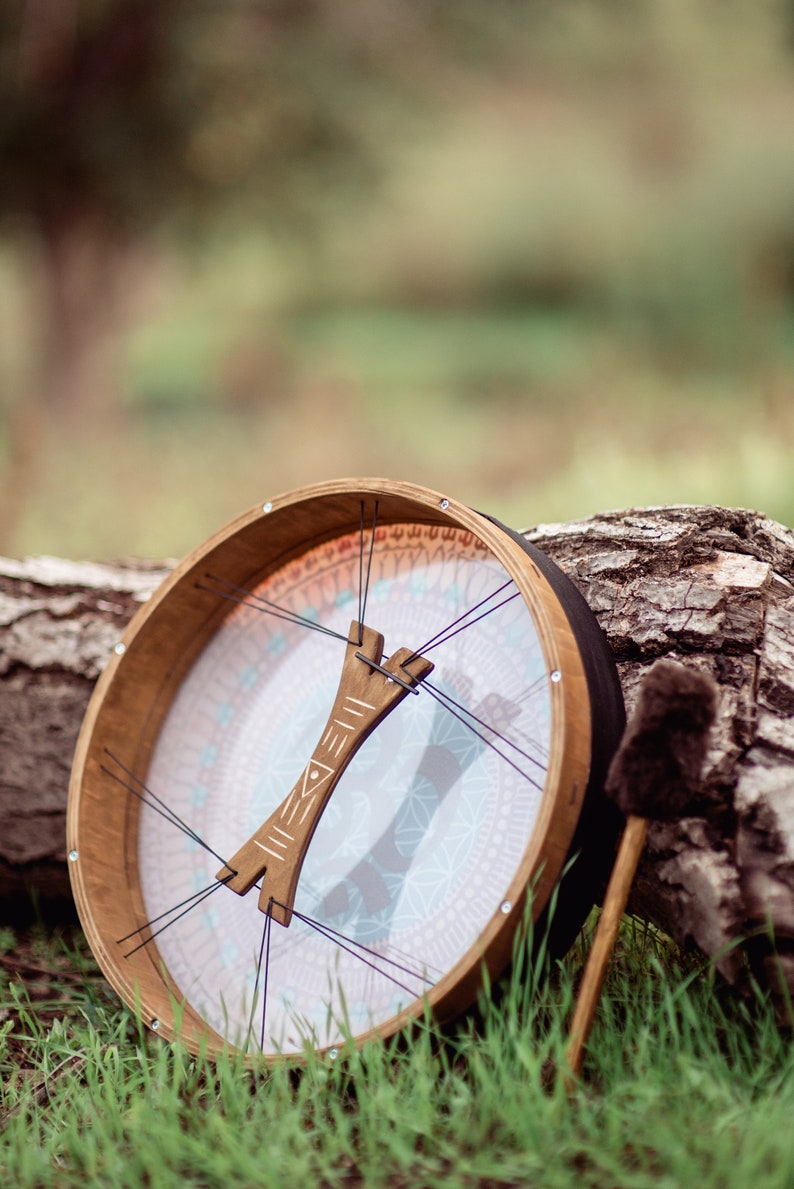 May include: A wooden frame drum with a white and blue patterned membrane and a wooden handle. The drum is resting on a tree trunk with a brown and black drum stick beside it.