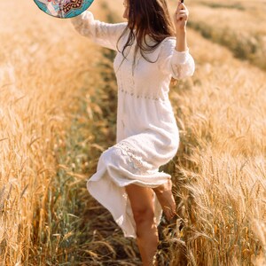 May include: A woman in a white dress dances in a field of wheat, holding a round, colourful drum with a mandala design and a black stick.