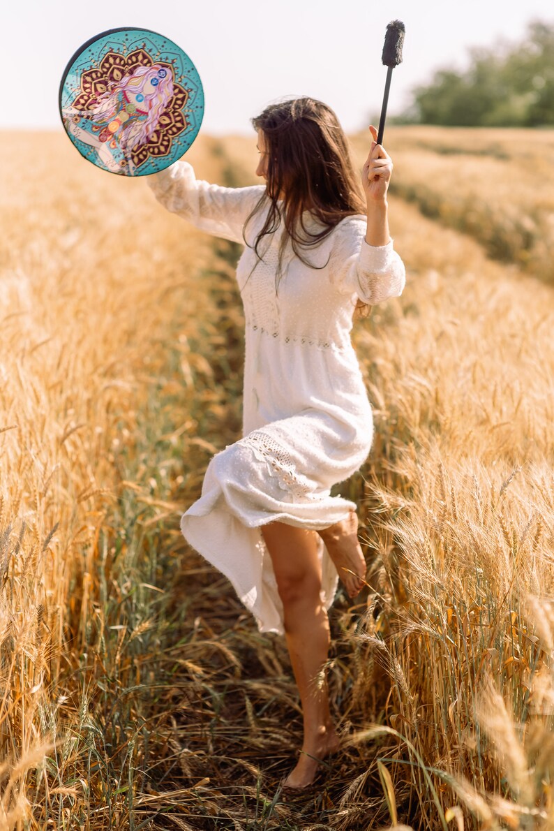 May include: A woman in a white dress dances in a field of wheat, holding a round, colourful drum with a mandala design and a black stick.