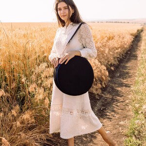 May include: A woman wearing a white lace dress stands in a field of wheat, holding a black round case with a strap.