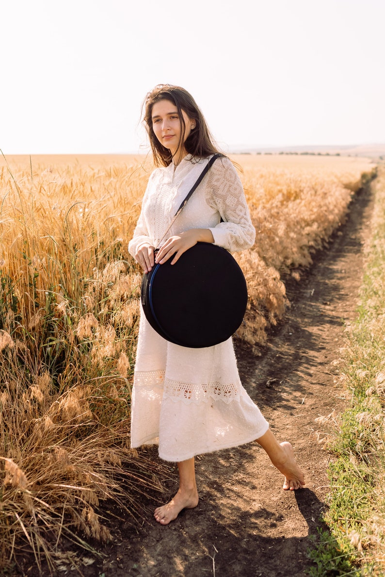 May include: A woman wearing a white lace dress stands in a field of wheat, holding a black round case with a strap.