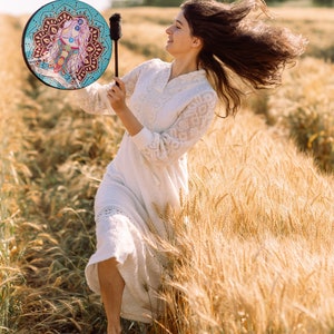 May include: A woman in a white dress plays a hand drum with a colourful mandala design in a field of wheat. Her hair is blowing in the wind.