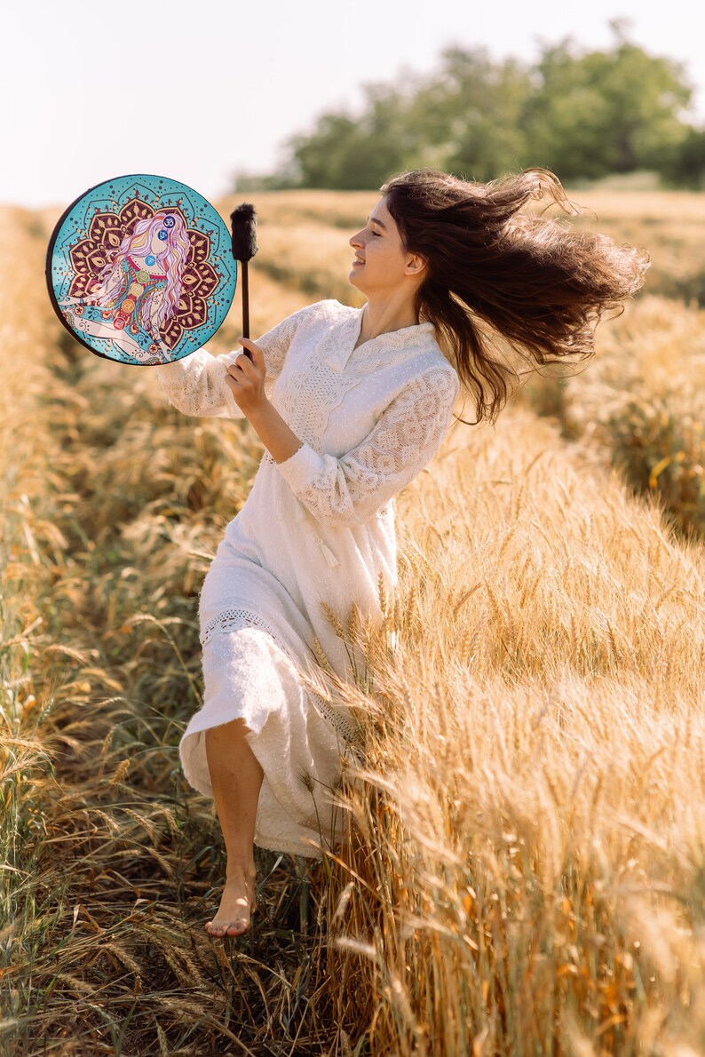 May include: A woman in a white dress plays a hand drum with a colourful mandala design in a field of wheat. Her hair is blowing in the wind.