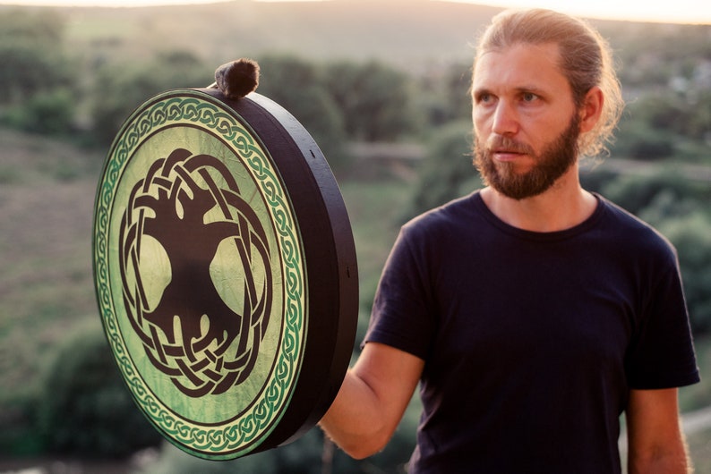 K&ouml;nnte beinhalten: Eine Person h&auml;lt eine runde Rahmtrommel aus Holz mit einem gr&uuml;nen und schwarzen keltischen Lebensbaum-Design. Die Trommel hat einen schwarzen Rand und eine braune Lederhaut.