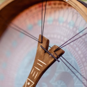 May include: Close-up of a wooden frame drum with a brown leather strap and black strings. The drum has a decorative design on the front with a geometric pattern in shades of blue, pink, and white.