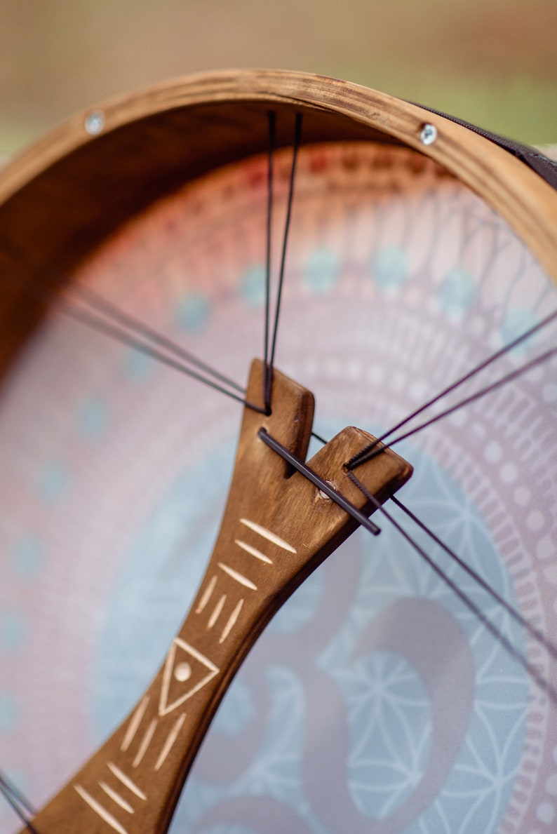 May include: Close-up of a wooden frame drum with a brown leather strap and black strings. The drum has a decorative design on the front with a geometric pattern in shades of blue, pink, and white.