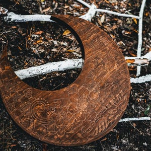 May include: A crescent-shaped wooden decorative piece with intricate floral and paisley carvings. The dark brown wood contrasts with the surrounding natural elements, including white branches and dark foliage. The carved design adds a touch of artistry.