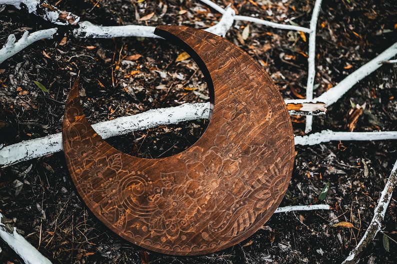 May include: A crescent-shaped wooden decorative piece with intricate floral and paisley carvings. The dark brown wood contrasts with the surrounding natural elements, including white branches and dark foliage. The carved design adds a touch of artistry.