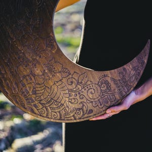 May include: A crescent moon-shaped wooden wall hanging with intricate floral and paisley carvings. The wood has a warm, brown tone, and the detailed patterns are etched into the surface. The design is held by a person wearing a black shirt.