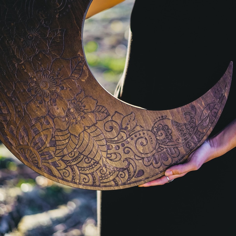 May include: A crescent moon-shaped wooden wall hanging with intricate floral and paisley carvings. The wood has a warm, brown tone, and the detailed patterns are etched into the surface. The design is held by a person wearing a black shirt.